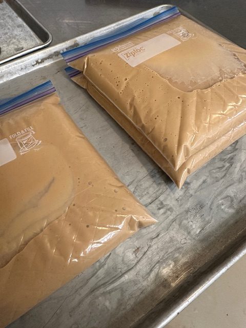 Two zip-top bags containing a smooth, creamy doughnut puree, placed on a metal baking tray.